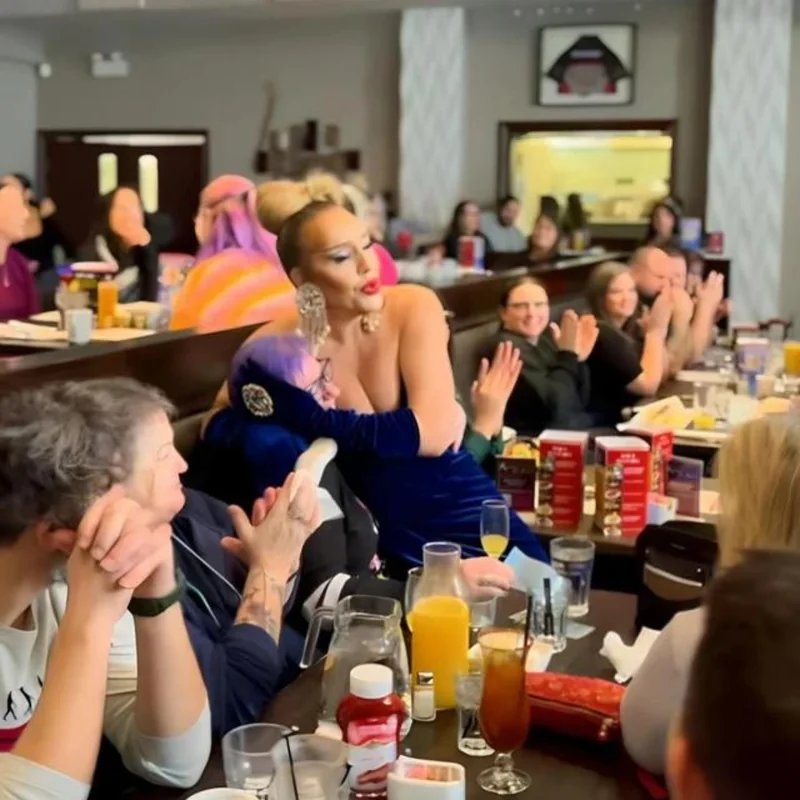 A drag performer leans into a table as brunch guests clap and cheer